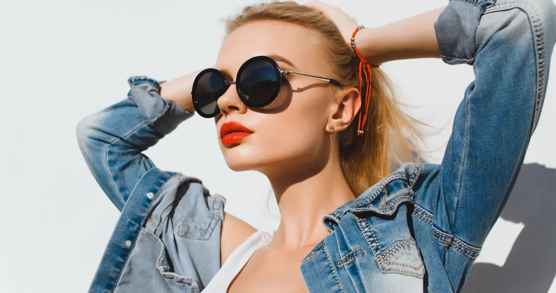Stylish blonde woman with ponytail, denim jacked over white tank top, black sunglasses, and red lipstick